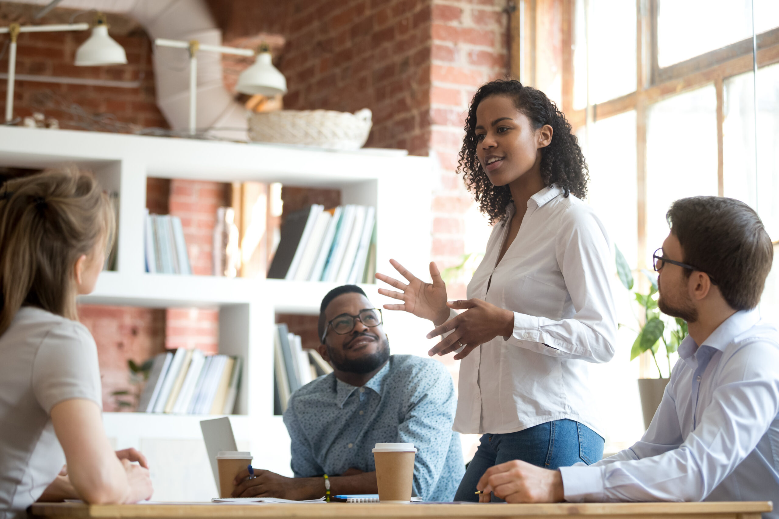 Black woman leader speaking to a group at a table with a bookshelf in the background